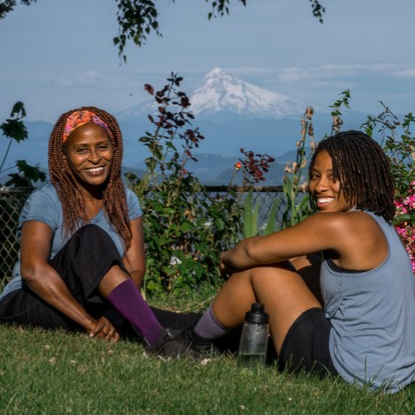A mother and daughter resk in the grass wearing TOughCutie socks with a beautiful mountain landscape behind them